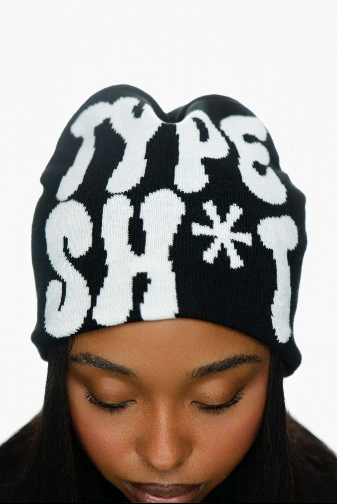 Black beanie with bold white text, worn by a person looking down, showing part of their face.