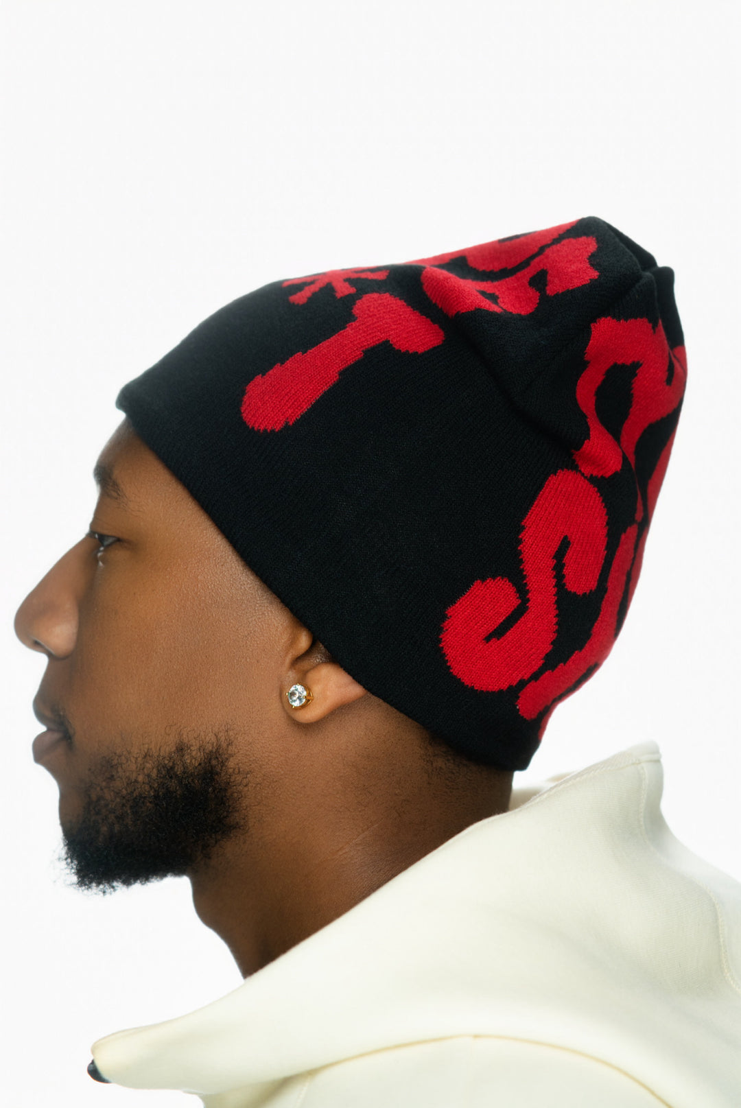Profile of a person wearing a black beanie with red designs, featuring an earring and light-colored jacket.
