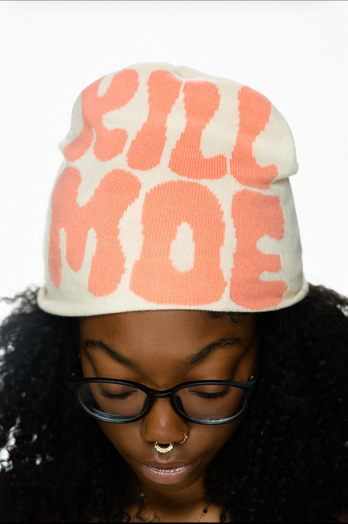 A person wearing a cream hat with pink text, glasses, and multiple nose piercings.