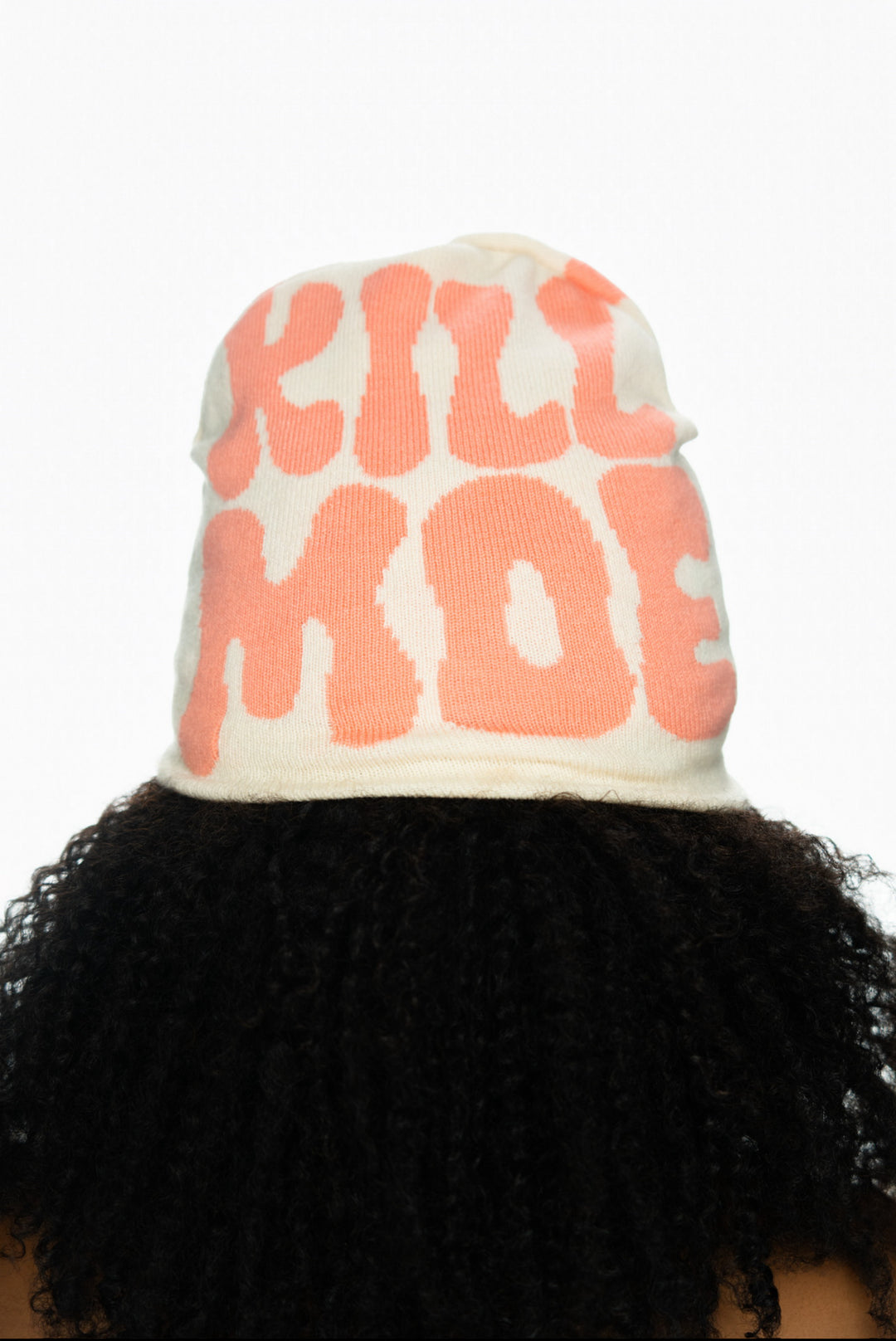 A beige beanie with bold pink text that reads "KILL MODE," worn on curly black hair.