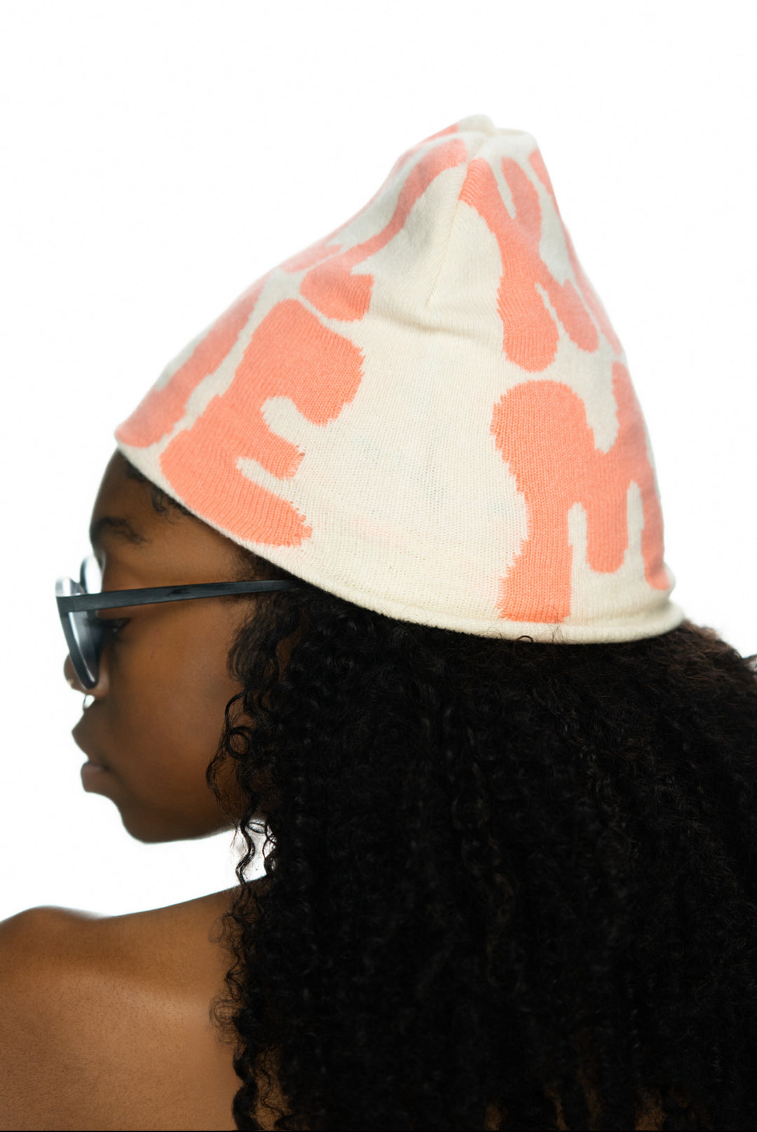 Person with curly hair wearing a light-colored beanie featuring coral patterns and glasses.