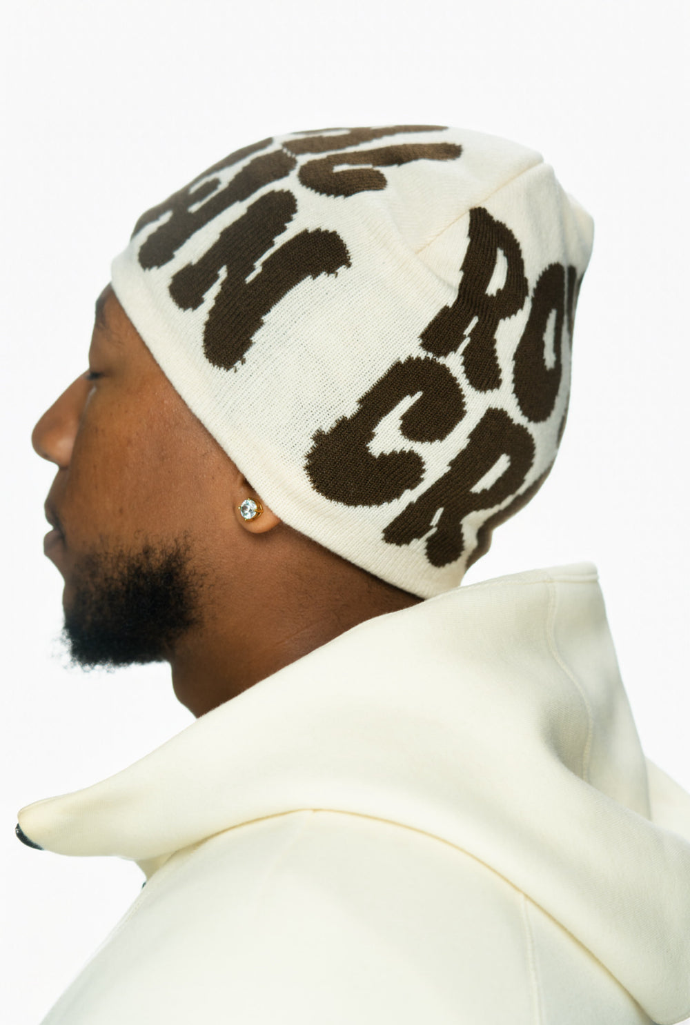 Profile view of a person wearing a beige beanie with dark brown lettering and a white hoodie.