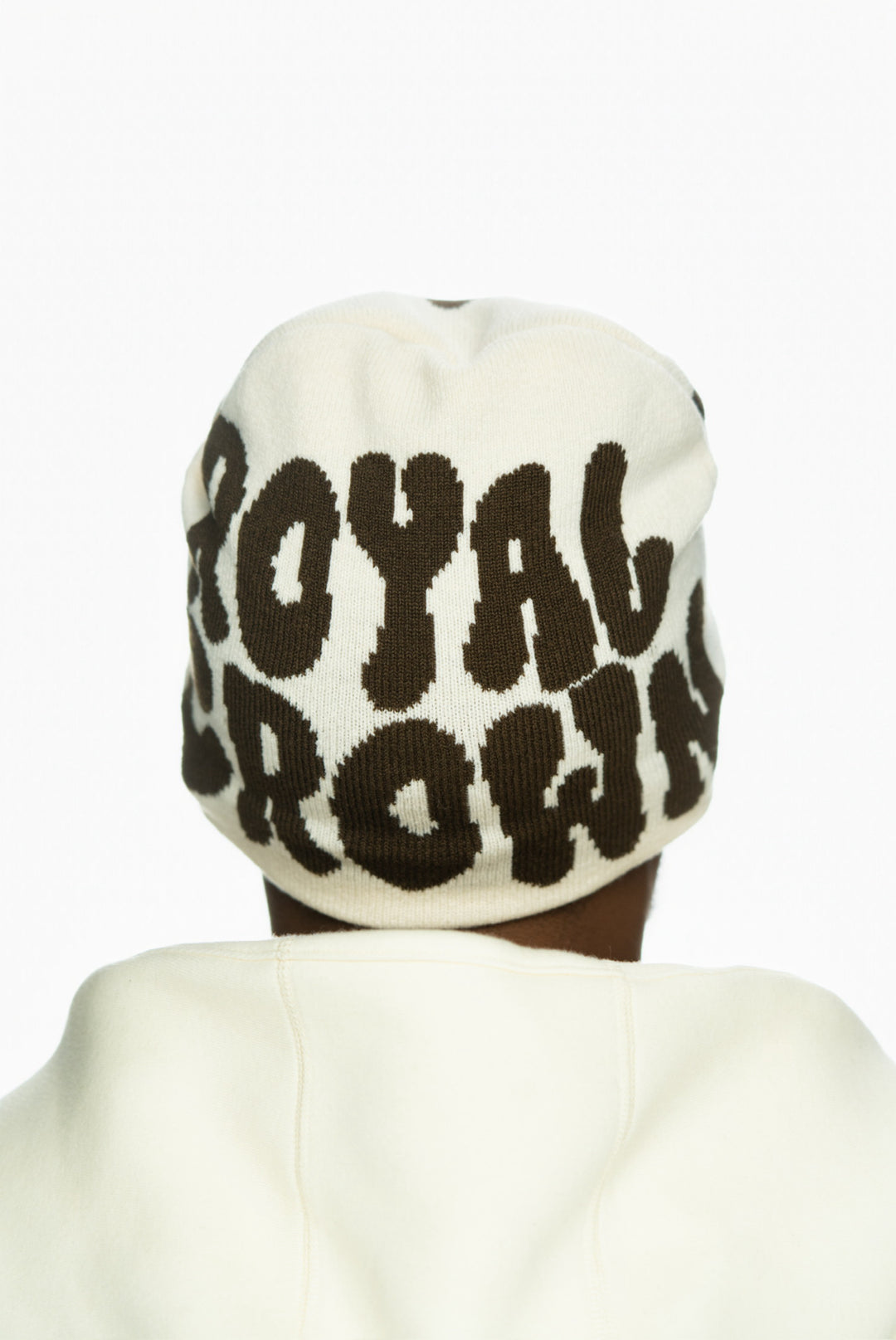 Cream beanie with bold brown lettering reading "ROYAL CROWN," worn by a person from behind.
