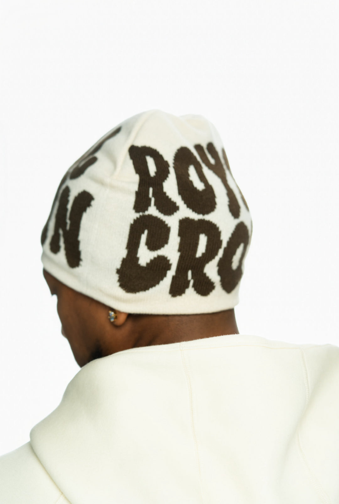 Cream beanie with brown text worn by a person, viewed from behind, with a white hoodie.