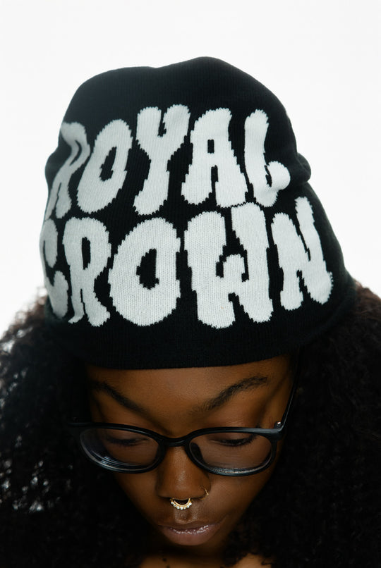 Black knit beanie with “ROYAL CROWN” in white letters worn by a person with curly hair and glasses.
