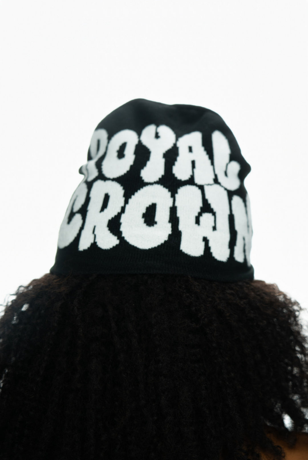 Black beanie with "ROYAL CROWN" text in bold white letters, worn on curly hair.