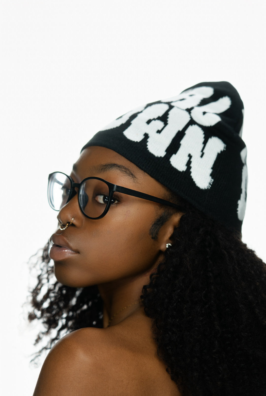 Young woman with curly hair, wearing glasses and a black beanie, poses against a white background.